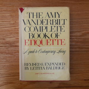 May include: A vintage book titled "The Amy Vanderbilt Complete Book of Etiquette." The cover is a light yellow with black and red text. The book is a guide to contemporary living, revised and expanded by Letitia Baldrige.
