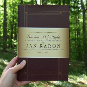 Peut inclure: Un livre à couverture rigide intitulé "Patches of Godlight" de Jan Karon. Le livre a une couverture marron foncé avec des accents dorés et une bande dorée au milieu avec le titre et le nom de l'auteur. Le livre est tenu devant un fond vert flou.