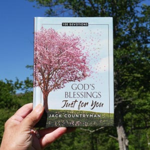 May include: A hardcover book titled "God's Blessings Just for You" by Jack Countryman. The cover features a pink flowering tree with the text "100 Devotions" at the top. The book is held against a backdrop of a blue sky and green foliage.