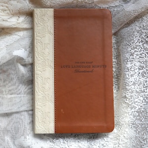 Op de afbeelding: Een bruin devotional boek met de tekst "THE ONE YEAR LOVE LANGUAGE MINUTE Devotional". Het boek heeft een crèmekleurige rug met een reliëf bloemmotief. Het boek is rechthoekig en lijkt van leer te zijn.