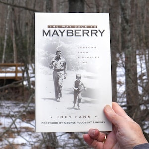 May include: A paperback book titled "THE WAY BACK TO MAYBERRY" is held up in front of a blurred outdoor background. The cover features a black and white image of a man and a child. The author is Joey Fann, with a foreword by George "Goober" Lindsey.