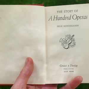 Może przedstawiać: Otwarta książka zatytułowana "The Story of A Hundred Operas" autorstwa Felixa Mendelssohna. Książka została wydana przez Grosset & Dunlap w Nowym Jorku.