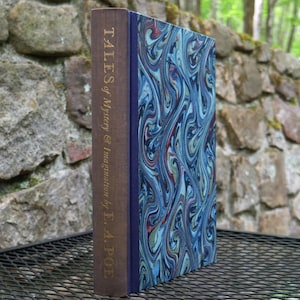 Könnte beinhalten: Ein gebundenes Buch mit dem Titel "Tales of Mystery & Imagination by E. A. Poe". Der Buchrücken ist braun mit goldener Schrift. Das Cover hat ein blaues, rotes und grünes Marmormuster. Das Buch steht auf einer Metalloberfläche.