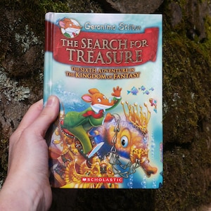 Könnte beinhalten: Kinderbuch "The Search for Treasure" von Geronimo Stilton. Das Cover zeigt eine Mausfigur, die auf einem fantastischen Unterwasserwesen reitet. Der Titel ist auf rotem Hintergrund geschrieben, mit dem Scholastic-Logo.