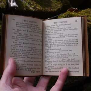 May include: An open, antique book with aged, yellowed pages. The text, titled "THE TEMPEST," is visible on both pages. The book is held open, revealing the text and the aged leather binding. The background is a natural, mossy setting.