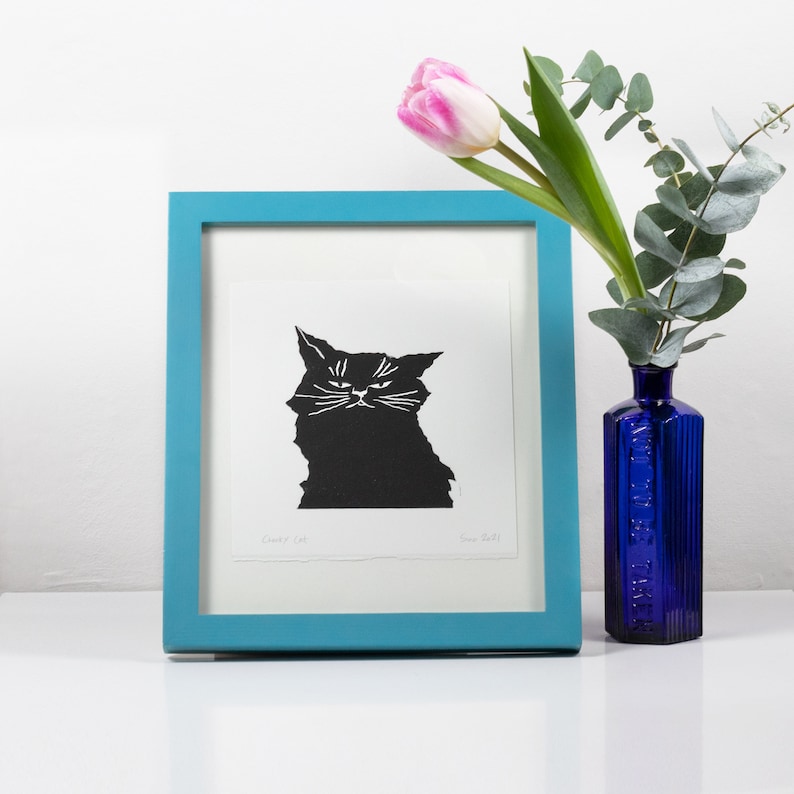 Puede incluir: Una impresi&oacute;n en blanco y negro enmarcada de un gato gru&ntilde;&oacute;n en un marco turquesa. Un tulip&aacute;n rosa y ramitas de eucalipto est&aacute;n en una botella azul cobalto. La impresi&oacute;n est&aacute; etiquetada como "Grumpy Cat" y firmada.