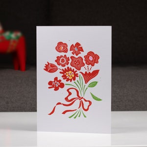 May include: A white card with a red and green floral design. The flowers are stylized and have a folk art feel. The bouquet is tied with a red ribbon.