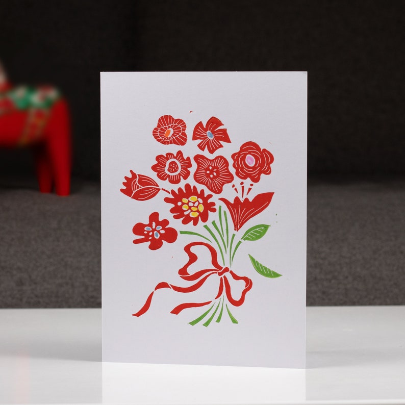May include: A white card with a red and green floral design. The flowers are stylized and have a folk art feel. The bouquet is tied with a red ribbon.