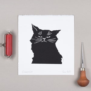 Puede incluir: Un grabado en lin&oacute;leo en blanco y negro de un gato gru&ntilde;&oacute;n. La impresi&oacute;n est&aacute; sobre papel blanco con el texto "Cranky Cat" y "Soo 2021". Un rodillo de impresi&oacute;n rojo y una herramienta de tallado son visibles.