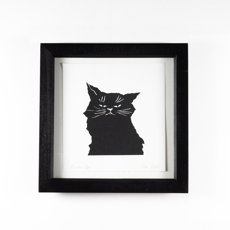 Puede incluir: Una impresi&oacute;n en blanco y negro enmarcada que presenta una ilustraci&oacute;n estilizada de un gato gru&ntilde;&oacute;n. El gato es una silueta negra con detalles blancos para los ojos y bigotes. La impresi&oacute;n est&aacute; enmarcada en un marco de madera negro. El texto "Grumpy Cat" es visible.