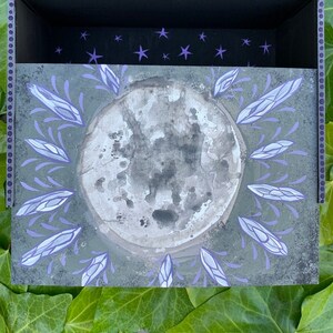 May include: A decorative box with a celestial theme. The box features a grey moon design surrounded by lavender accents. The interior is black with purple star details. The box is set against a backdrop of green leaves.