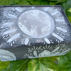 Puede incluir: Una caja rectangular de madera gris oscuro con un dise&ntilde;o celestial. La tapa presenta una gran luna gris texturizada rodeada de formas blancas en forma de cristal. Los lados est&aacute;n adornados con motivos similares de cristal y estrellas.