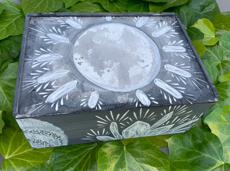 Puede incluir: Una caja rectangular de madera gris oscuro con un dise&ntilde;o celestial. La tapa presenta una gran luna gris texturizada rodeada de formas blancas en forma de cristal. Los lados est&aacute;n adornados con motivos similares de cristal y estrellas.