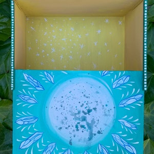 Op de afbeelding: Een handgeschilderde houten kist met een hemels thema. De buitenkant is turquoise met een grote maan en kristalontwerpen. De binnenkant is geel met witte sterpatronen. De doos heeft een gestippelde rand.