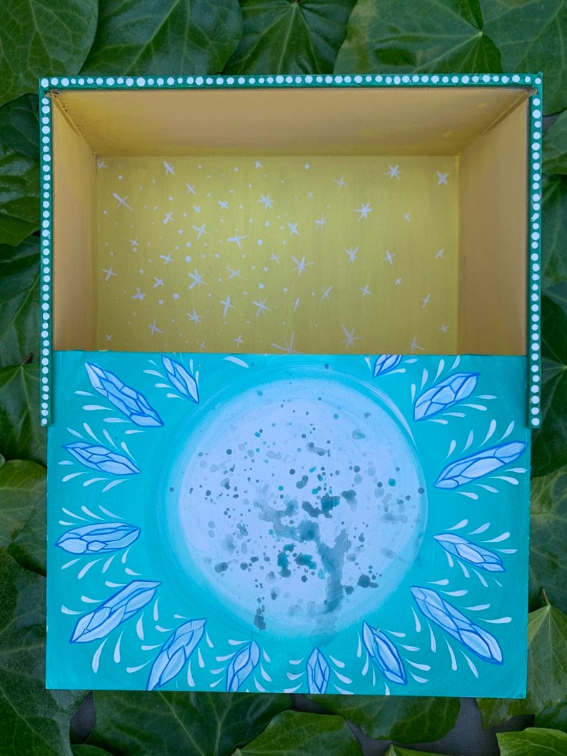 Op de afbeelding: Een handgeschilderde houten kist met een hemels thema. De buitenkant is turquoise met een grote maan en kristalontwerpen. De binnenkant is geel met witte sterpatronen. De doos heeft een gestippelde rand.