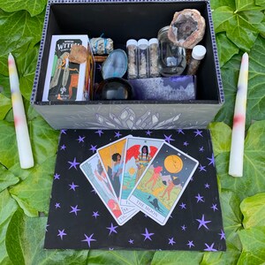 May include: An open black box with a crystal design, holding a tarot deck, crystals, and jars. Four tarot cards are on a black surface with purple stars. Two tall, multi-coloured candles are positioned beside the box. The box contains various spiritual items.