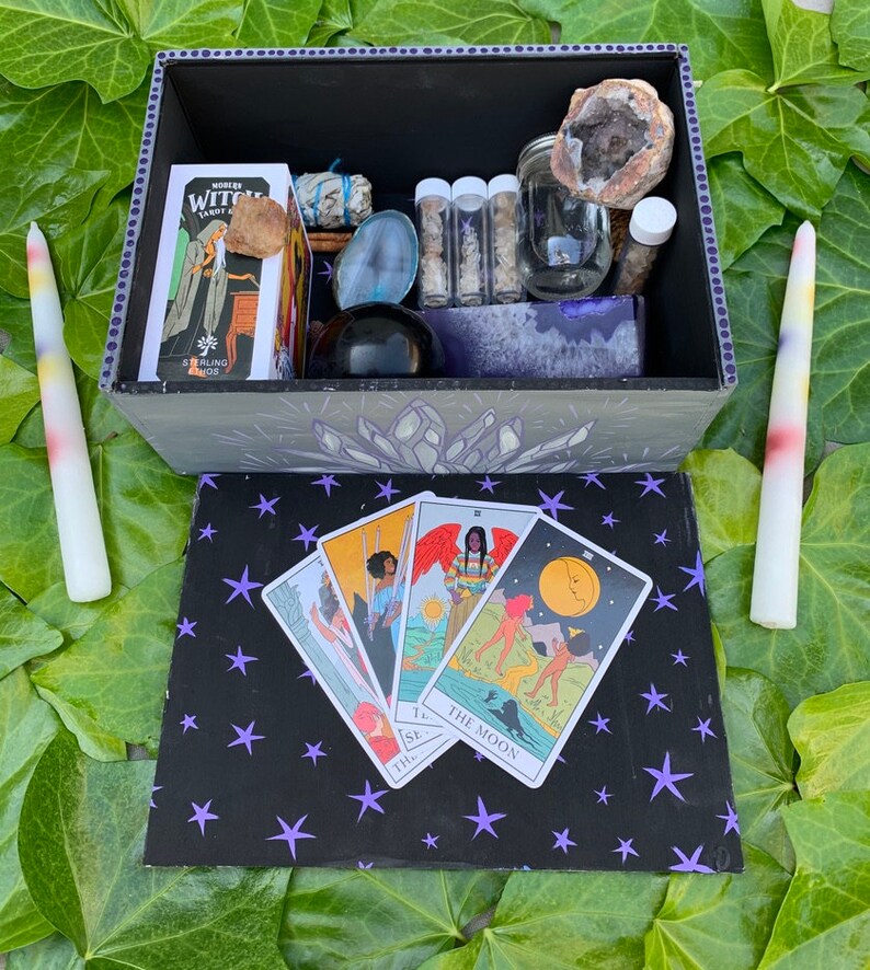 May include: An open black box with a crystal design, holding a tarot deck, crystals, and jars. Four tarot cards are on a black surface with purple stars. Two tall, multi-coloured candles are positioned beside the box. The box contains various spiritual items.