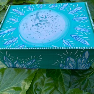 Op de afbeelding: Een rechthoekige houten doos, turquoise geschilderd met een hemels ontwerp. Het deksel heeft een grote, lichtblauwe cirkel met een spat-effect, omringd door blauwe kristalachtige vormen. De zijkanten hebben vergelijkbare kristalontwerpen.