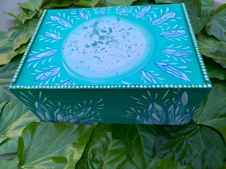 Op de afbeelding: Een rechthoekige houten doos, turquoise geschilderd met een hemels ontwerp. Het deksel heeft een grote, lichtblauwe cirkel met een spat-effect, omringd door blauwe kristalachtige vormen. De zijkanten hebben vergelijkbare kristalontwerpen.