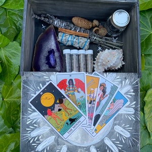 Puede incluir: Una caja de madera gris con cartas del tarot, una geoda morada, manojos de salvia, una concha y peque&ntilde;os viales. La caja est&aacute; rodeada de hojas verdes. Las cartas del tarot son visibles.