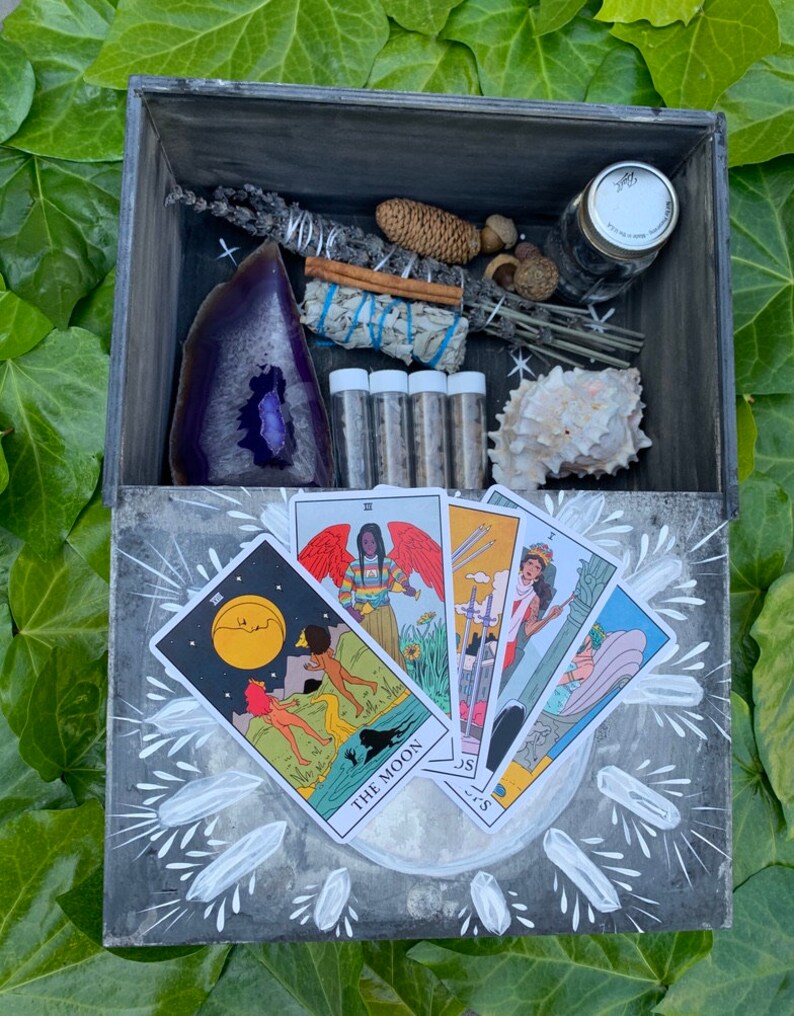 Puede incluir: Una caja de madera gris con cartas del tarot, una geoda morada, manojos de salvia, una concha y peque&ntilde;os viales. La caja est&aacute; rodeada de hojas verdes. Las cartas del tarot son visibles.