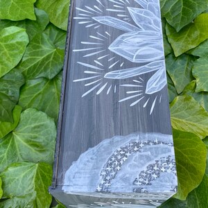 Puede incluir: Una caja de madera gris con un dise&ntilde;o de cristal blanco y rayos de estrellas. La caja tiene una tapa con bisagras y est&aacute; decorada con pintura blanca. La caja est&aacute; sobre un fondo de hojas verdes.