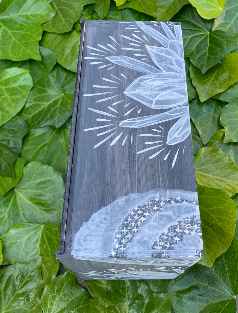Puede incluir: Una caja de madera gris con un dise&ntilde;o de cristal blanco y rayos de estrellas. La caja tiene una tapa con bisagras y est&aacute; decorada con pintura blanca. La caja est&aacute; sobre un fondo de hojas verdes.
