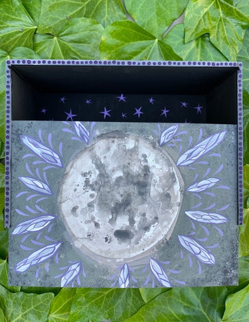 May include: A decorative box with a celestial theme. The box features a grey moon design surrounded by lavender accents. The interior is black with purple star details. The box is set against a backdrop of green leaves.