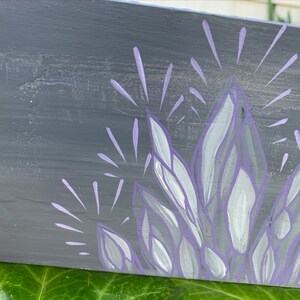 May include: A hand-painted wooden sign with a grey background and a cluster of crystals in shades of purple and white. The crystals are stylized with radiating lines, creating a sparkling effect. The sign is resting on green leaves.