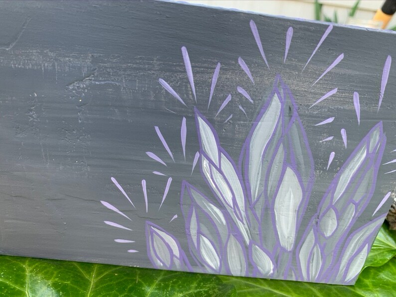 May include: A hand-painted wooden sign with a grey background and a cluster of crystals in shades of purple and white. The crystals are stylized with radiating lines, creating a sparkling effect. The sign is resting on green leaves.