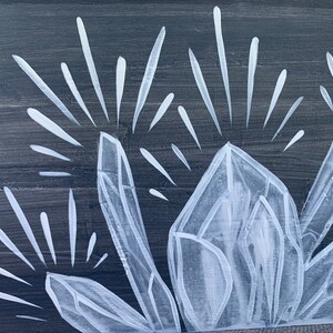 Puede incluir: Primer plano de un panel de madera pintado con un dise&ntilde;o de grupo de cristales blancos. Los cristales est&aacute;n detallados con facetas geom&eacute;tricas y l&iacute;neas radiantes, sobre un fondo gris oscuro. La obra evoca un tema natural y espiritual.