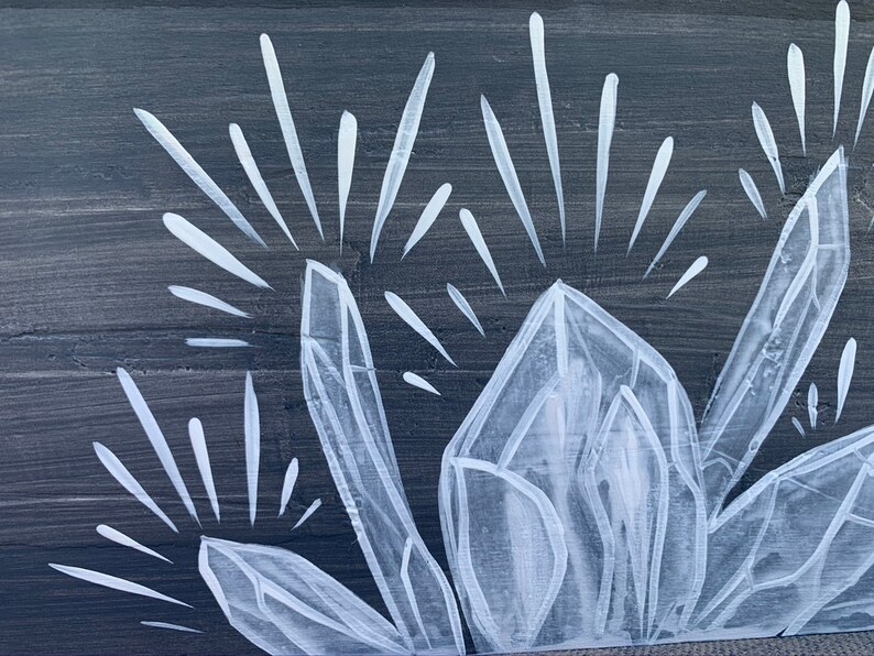 Puede incluir: Primer plano de un panel de madera pintado con un dise&ntilde;o de grupo de cristales blancos. Los cristales est&aacute;n detallados con facetas geom&eacute;tricas y l&iacute;neas radiantes, sobre un fondo gris oscuro. La obra evoca un tema natural y espiritual.