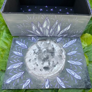 May include: A hand-painted wooden box in grey, with a lid decorated with celestial and crystal designs. The lid shows a full moon encircled by crystal-like shapes in grey and purple. The box interior is black with purple star accents.