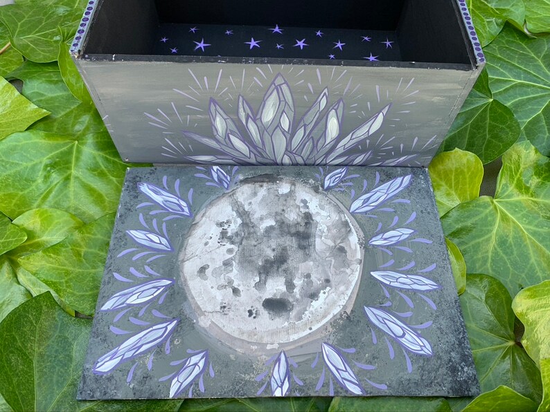 May include: A hand-painted wooden box in grey, with a lid decorated with celestial and crystal designs. The lid shows a full moon encircled by crystal-like shapes in grey and purple. The box interior is black with purple star accents.