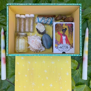 Op de afbeelding: Een gele doos met diverse items, waaronder een tarotdeck, kristallen, een zeeschelp en kleine glazen flesjes. Twee lange, kleurrijke kaarsen flankeren de doos. De doos is versierd met witte stippen en een geel sterrenpatroon.
