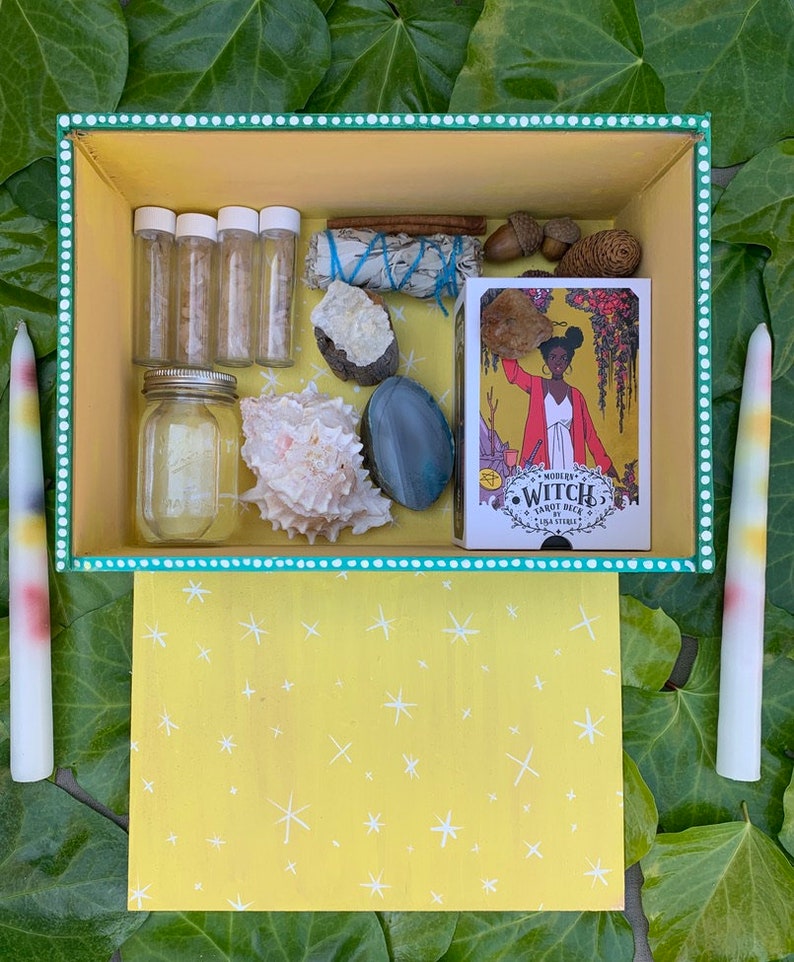 Op de afbeelding: Een gele doos met diverse items, waaronder een tarotdeck, kristallen, een zeeschelp en kleine glazen flesjes. Twee lange, kleurrijke kaarsen flankeren de doos. De doos is versierd met witte stippen en een geel sterrenpatroon.