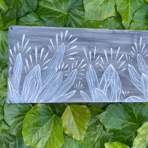 Puede incluir: Un panel rectangular de madera pintado de gris con un dise&ntilde;o de cristales blancos. Los cristales est&aacute;n estilizados con bordes afilados y l&iacute;neas radiantes, sobre un fondo gris texturizado. La obra de arte se exhibe sobre un lecho de hiedra verde.