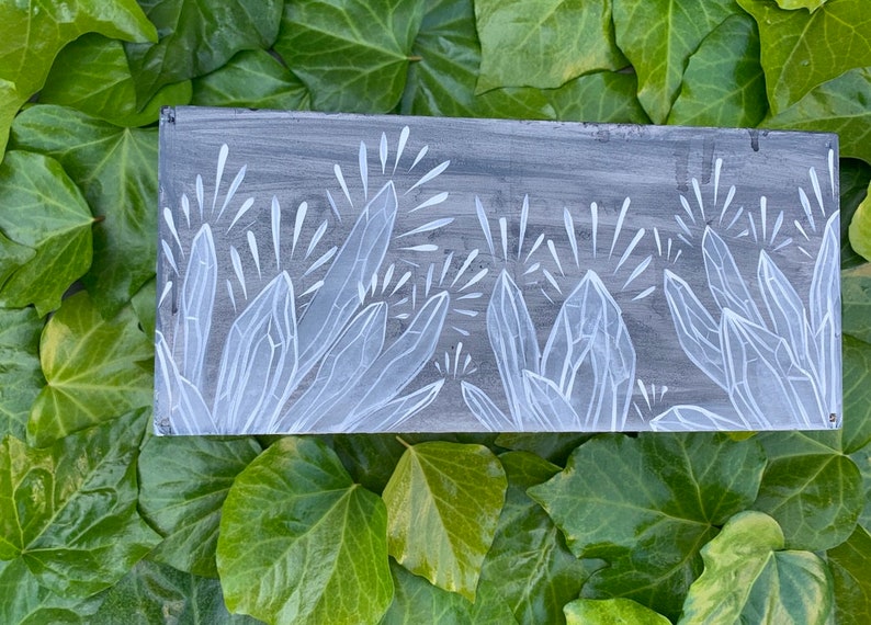 Puede incluir: Un panel rectangular de madera pintado de gris con un dise&ntilde;o de cristales blancos. Los cristales est&aacute;n estilizados con bordes afilados y l&iacute;neas radiantes, sobre un fondo gris texturizado. La obra de arte se exhibe sobre un lecho de hiedra verde.