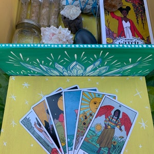 Op de afbeelding: Een houten kist met een groen en wit bloemmotief, gevuld met diverse items waaronder glazen flesjes, een pot, kristallen en een tarotkaartendeck. De tarotkaarten zijn uitgespreid op een gele ondergrond, met kleurrijke illustraties.
