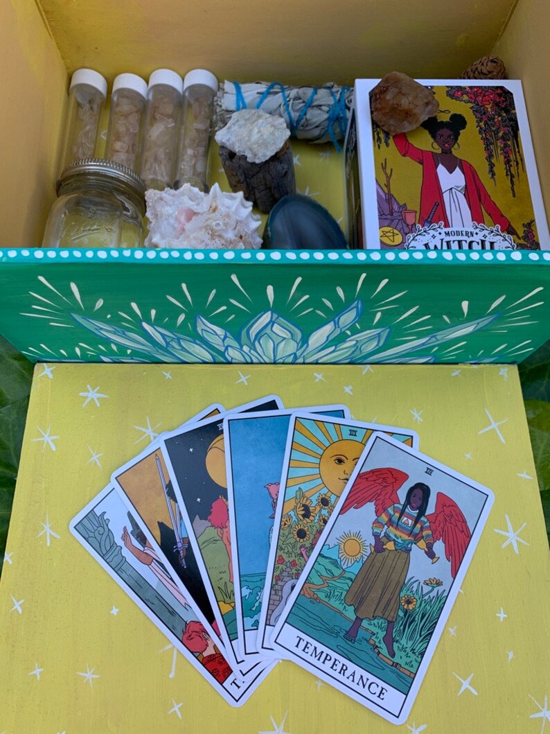 Op de afbeelding: Een houten kist met een groen en wit bloemmotief, gevuld met diverse items waaronder glazen flesjes, een pot, kristallen en een tarotkaartendeck. De tarotkaarten zijn uitgespreid op een gele ondergrond, met kleurrijke illustraties.
