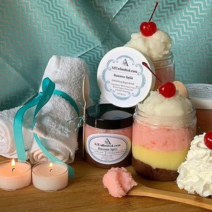May include: A set of three bath and body products with a banana split theme. The products are in glass jars with labels that say "SJ's Unlimited.com Banana Split Exfoliating Sugar Scrub". The jars are arranged on a wooden surface with a white towel, two candles, and a wooden spoon.