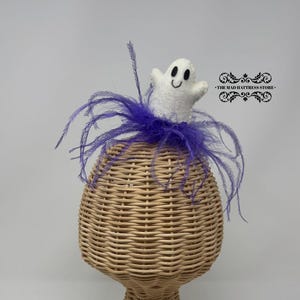 May include: A whimsical Halloween-themed hat, featuring a woven wicker base and a plush white ghost with black eyes and a smiling mouth. The ghost is adorned with purple feathers. The Mad Hatteress Store logo is visible.