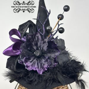 May include: A black felt witch hat featuring a large purple and black flower, a purple satin ribbon bow, black feathers, and decorative black berry sprigs. The text THE MAD HATTRESS STORE is printed in black on the upper left side of the image.