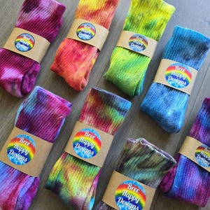 Peut inclure: Huit paires de chaussettes montantes c&ocirc;tel&eacute;es avec divers motifs tie-dye aux couleurs rose, orange, jaune, vert et bleu. Chaque paire est maintenue par une bande en papier brun portant un logo circulaire indiquant Bree Happy Designs.