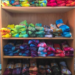 Peut inclure: Plusieurs piles de chaussettes tie-dye dispos&eacute;es sur des &eacute;tag&egrave;res en bois. Les chaussettes pr&eacute;sentent diverses combinaisons de couleurs, notamment des jaunes, verts, bleus, roses, rouges et violets vifs. Des bandes en papier beige les maintiennent.