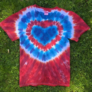 May include: A tie-dye V-neck t-shirt with a heart design in red, white, and blue. The heart shape is centered on the chest, with the colors radiating outwards. The shirt is made of a soft material and is laying on a bed of green grass.