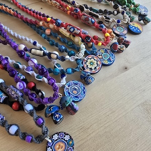 May include: A collection of handmade macrame necklaces with colorful beads and decorative pendants. The necklaces feature various colors, including red, blue, purple, and green, with unique pendant designs.