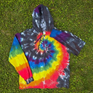 May include: A tie-dye zip-up hoodie with a spiral design in vibrant rainbow colours. The hoodie features a black hood and sleeves, and a full-length zip. The background is green grass.