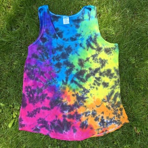 May include: A sleeveless tie-dye vest top in a vibrant rainbow of colours, including blue, purple, pink, yellow, and orange. The fabric has a swirling pattern with dark accents. The vest top is displayed on green grass.