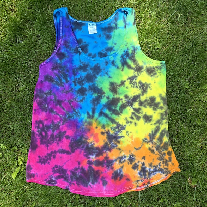 May include: A sleeveless tie-dye vest top in a vibrant rainbow of colours, including blue, purple, pink, yellow, and orange. The fabric has a swirling pattern with dark accents. The vest top is displayed on green grass.